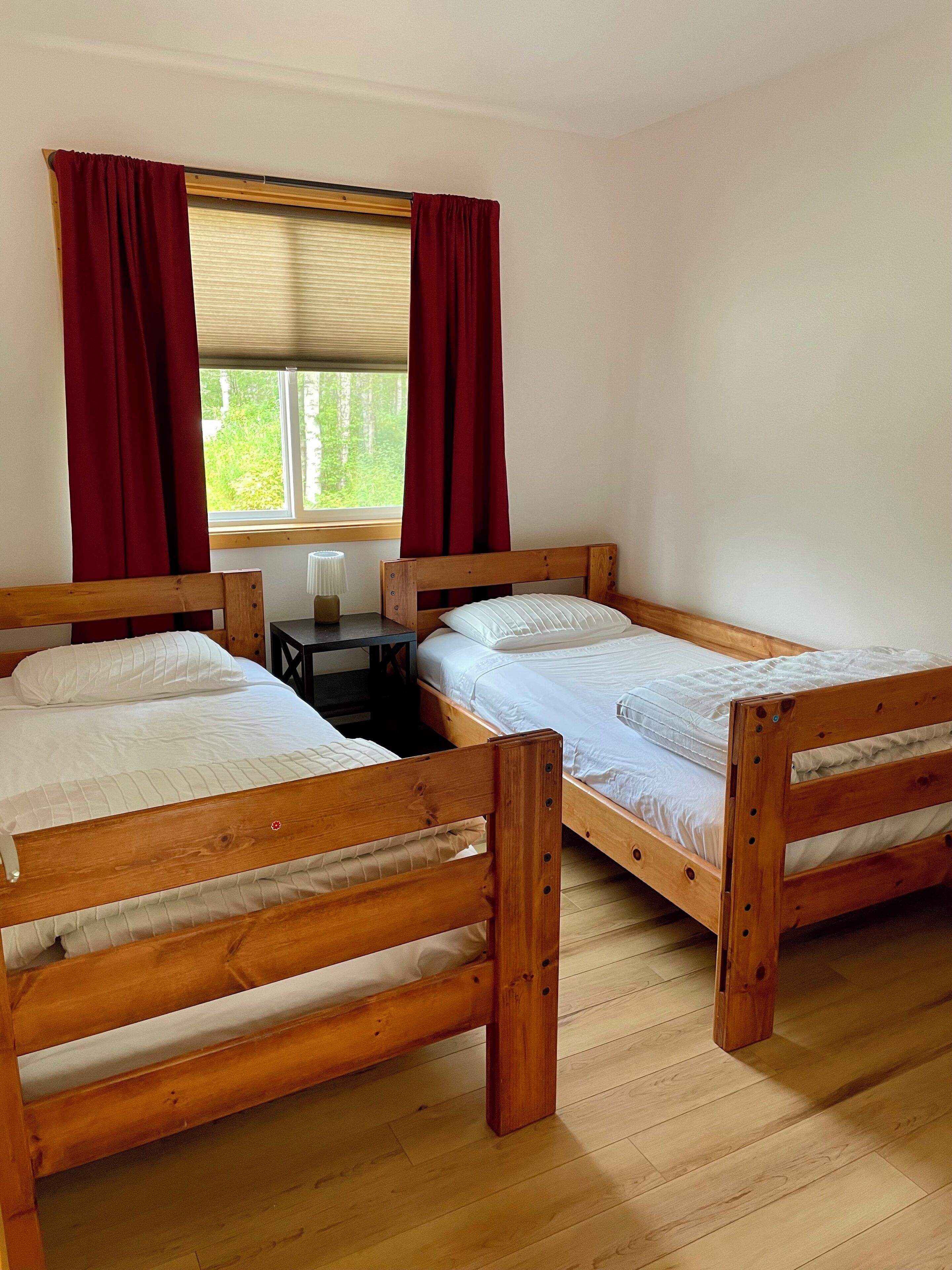 A room with two wooden beds, a small table, and a window with red curtains.