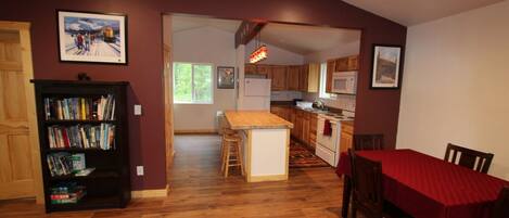A spacious living room with a wooden floor, a dining area with a red tablecloth, and a kitchen with wooden cabinets.
