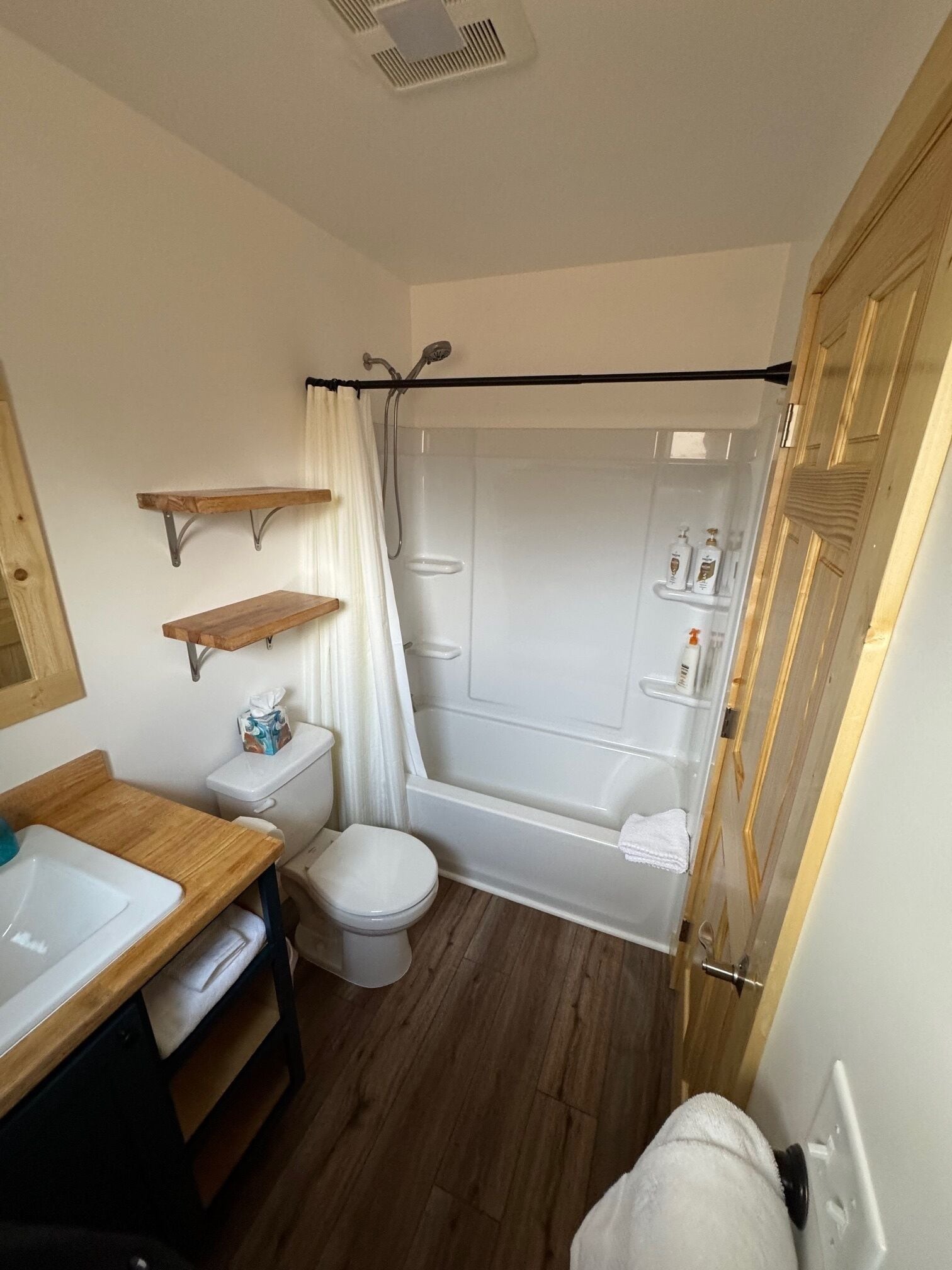 A compact bathroom with a toilet, sink, and bathtub. There are wooden shelves above the toilet and a shower curtain over the bathtub.