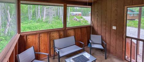 Une cabane avec un intérieur en bois, un ventilateur de plafond, une table avec des chaises et de grandes fenêtres offrant une vue sur une forêt et une autre cabane au loin.