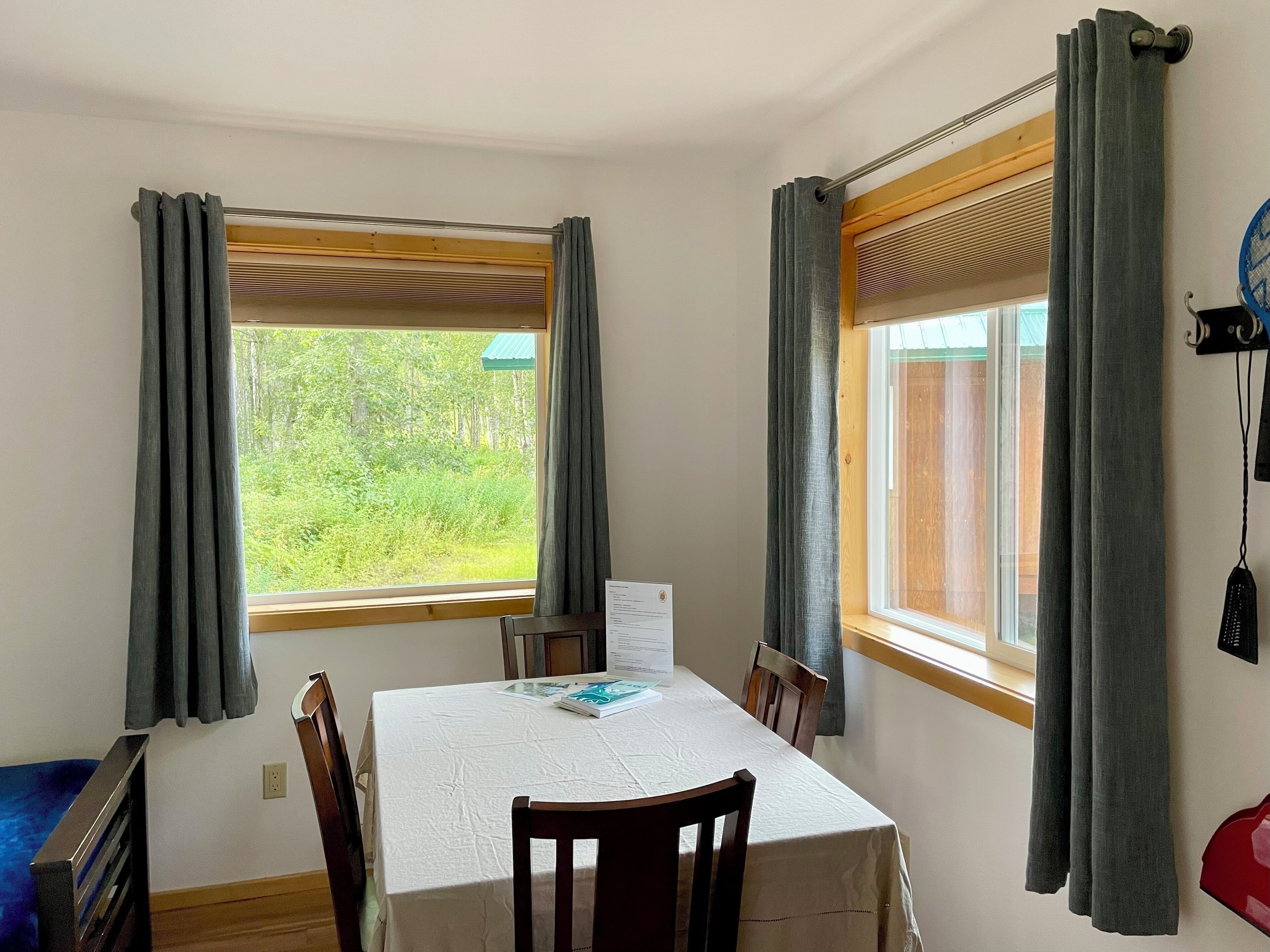 A dining area with a table set for two, two chairs, a window with curtains, and a view of greenery outside.