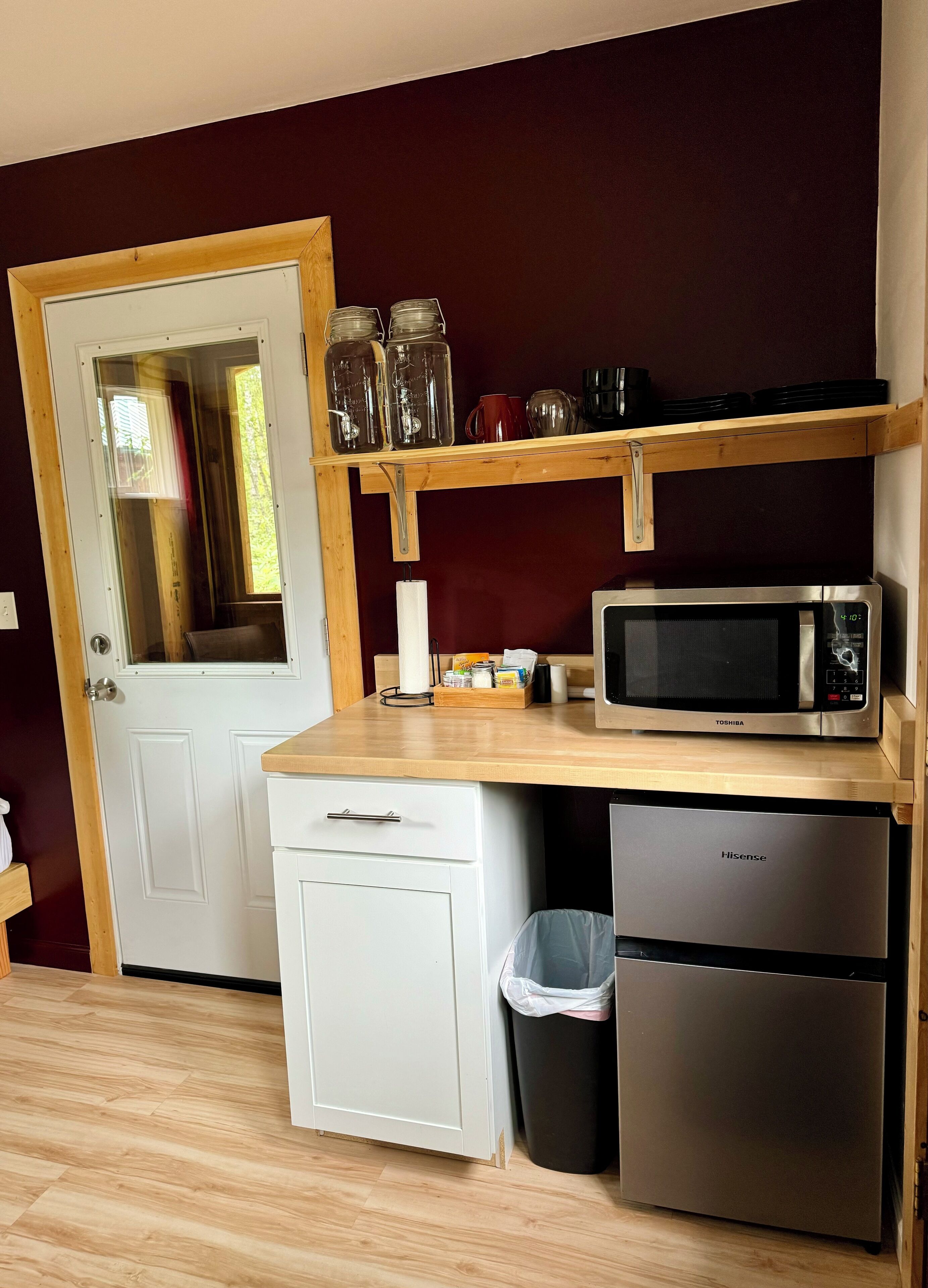 A compact kitchen with a microwave, a small refrigerator, a trash bin, and a wooden shelf with jars and mugs.