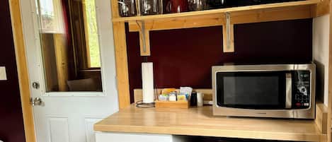 A compact kitchen with a microwave, a small refrigerator, a trash bin, and a wooden shelf with jars and mugs.