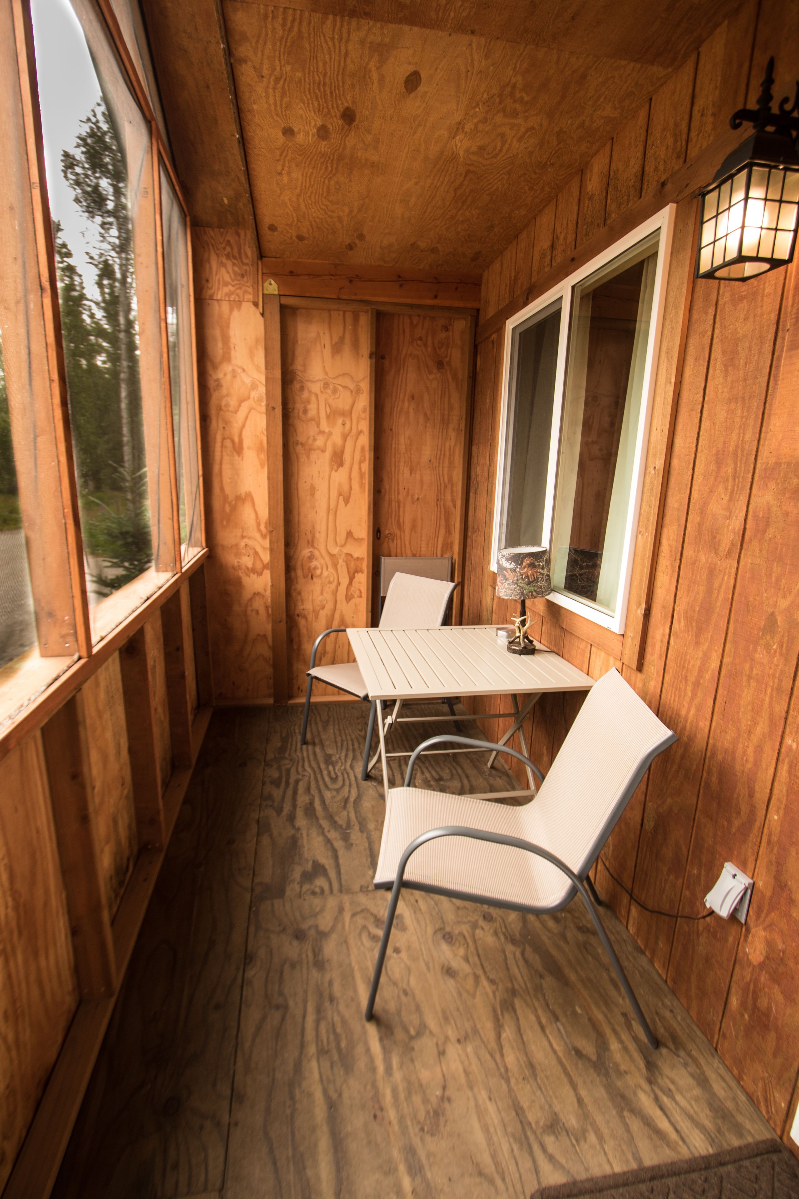 A wooden interior with a table and two chairs, large windows, and a wall-mounted light fixture.