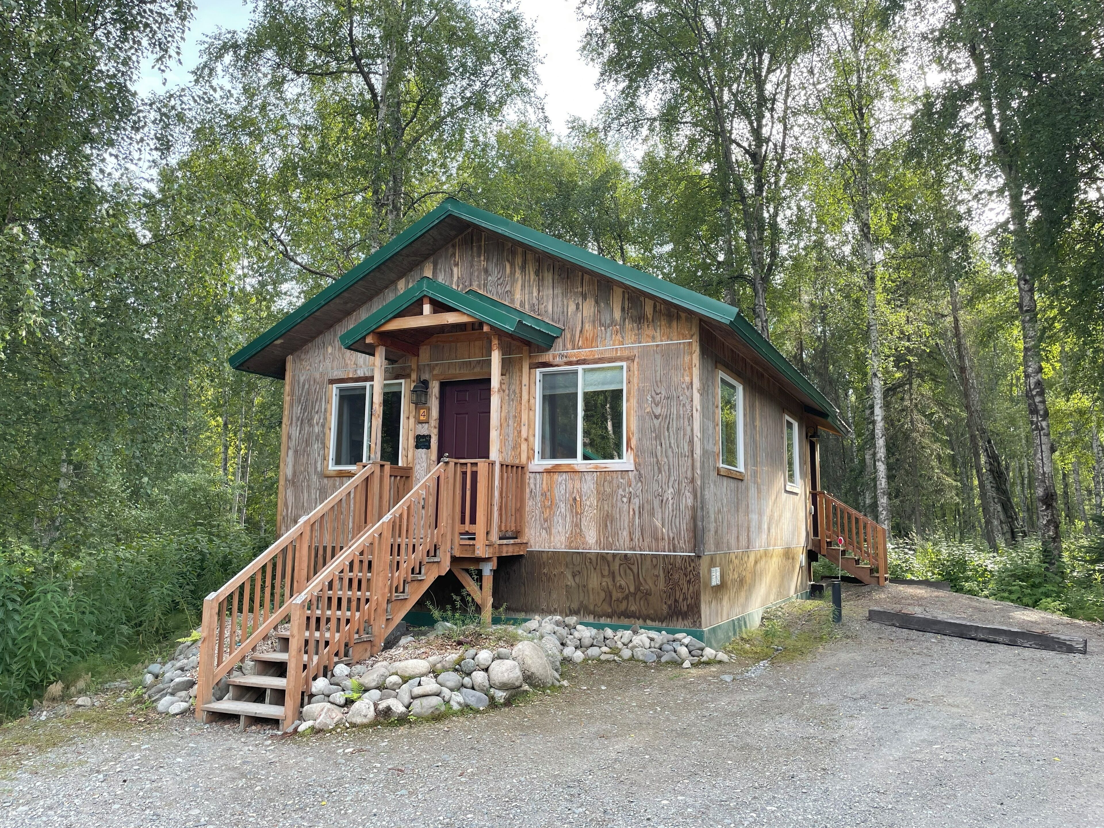 A wooden cabin with a green roof, a front porch with a wooden staircase, and a gravel driveway.