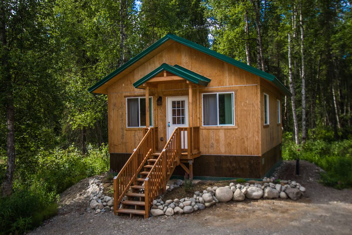 A wooden cabin with a green roof, a front door, and a staircase leading to the entrance.