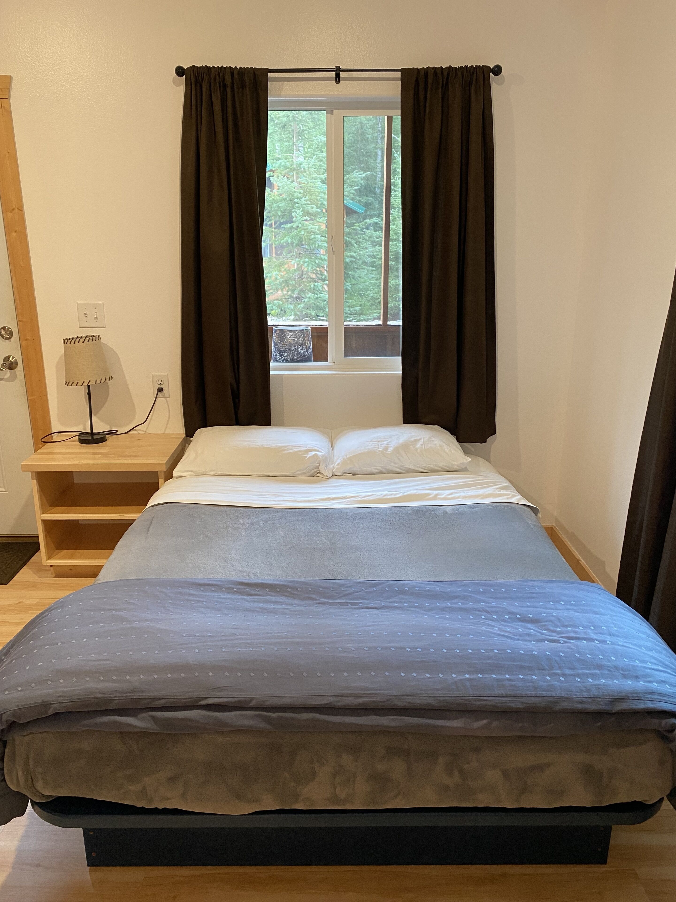 Una cama bien hecha con edredón azul y blanco, una lámpara de noche y una ventana con cortinas.