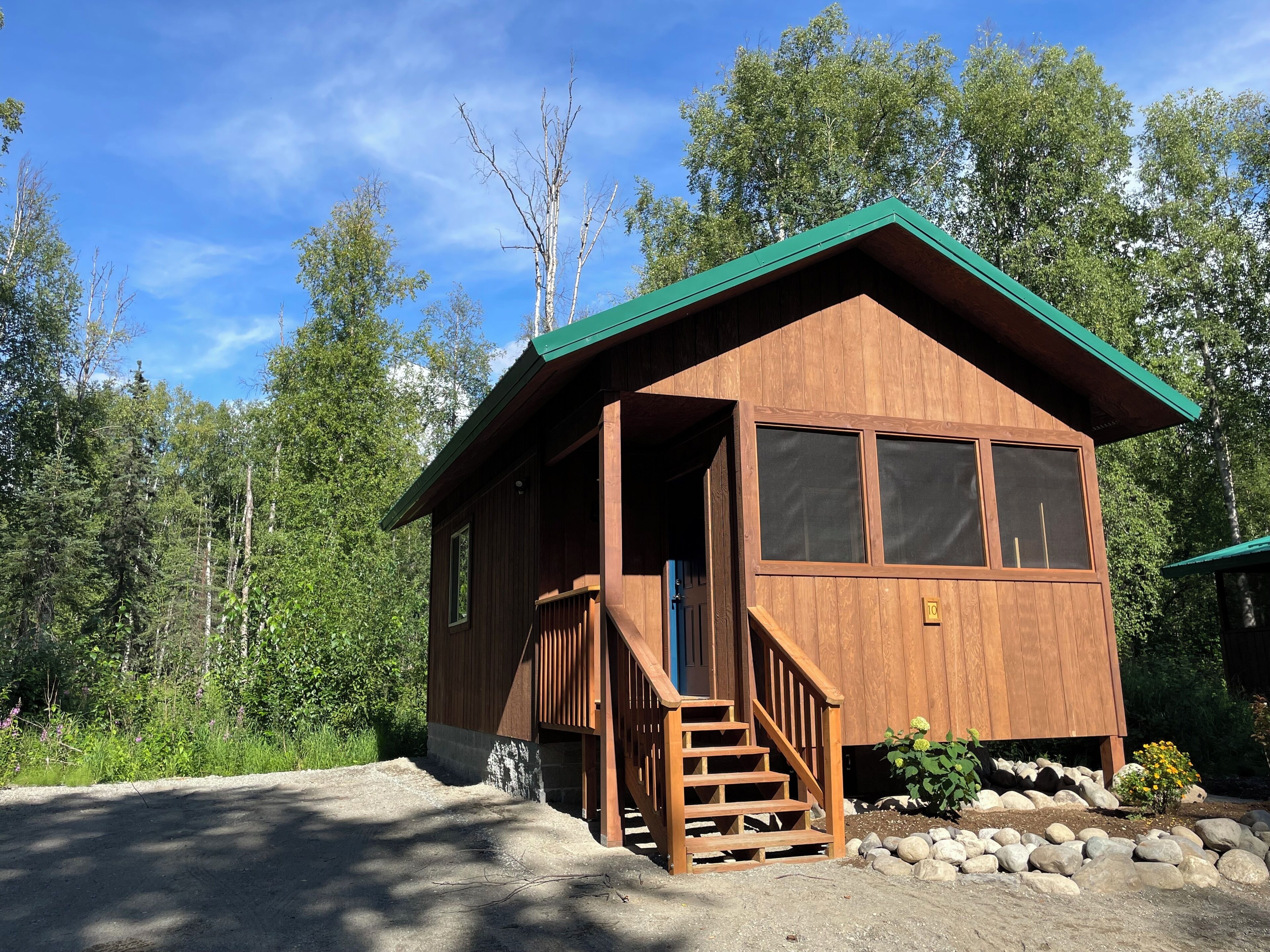 A wooden cabin with a green roof, a front porch, and a stone foundation.