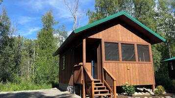 A wooden cabin with a green roof, a front porch, and a stone foundation.