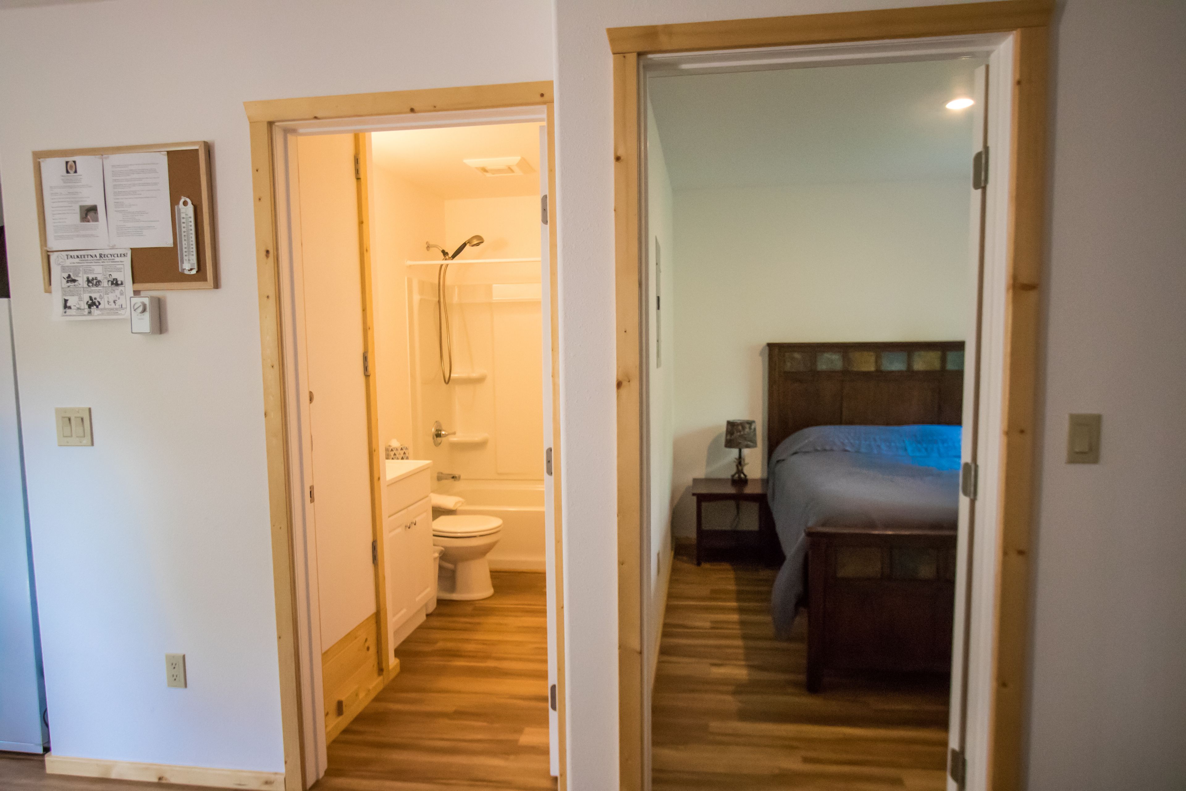 Una habitación con piso de madera, una cama con cubrecama azul, una mesita de noche con lámpara y un cabecero de madera.