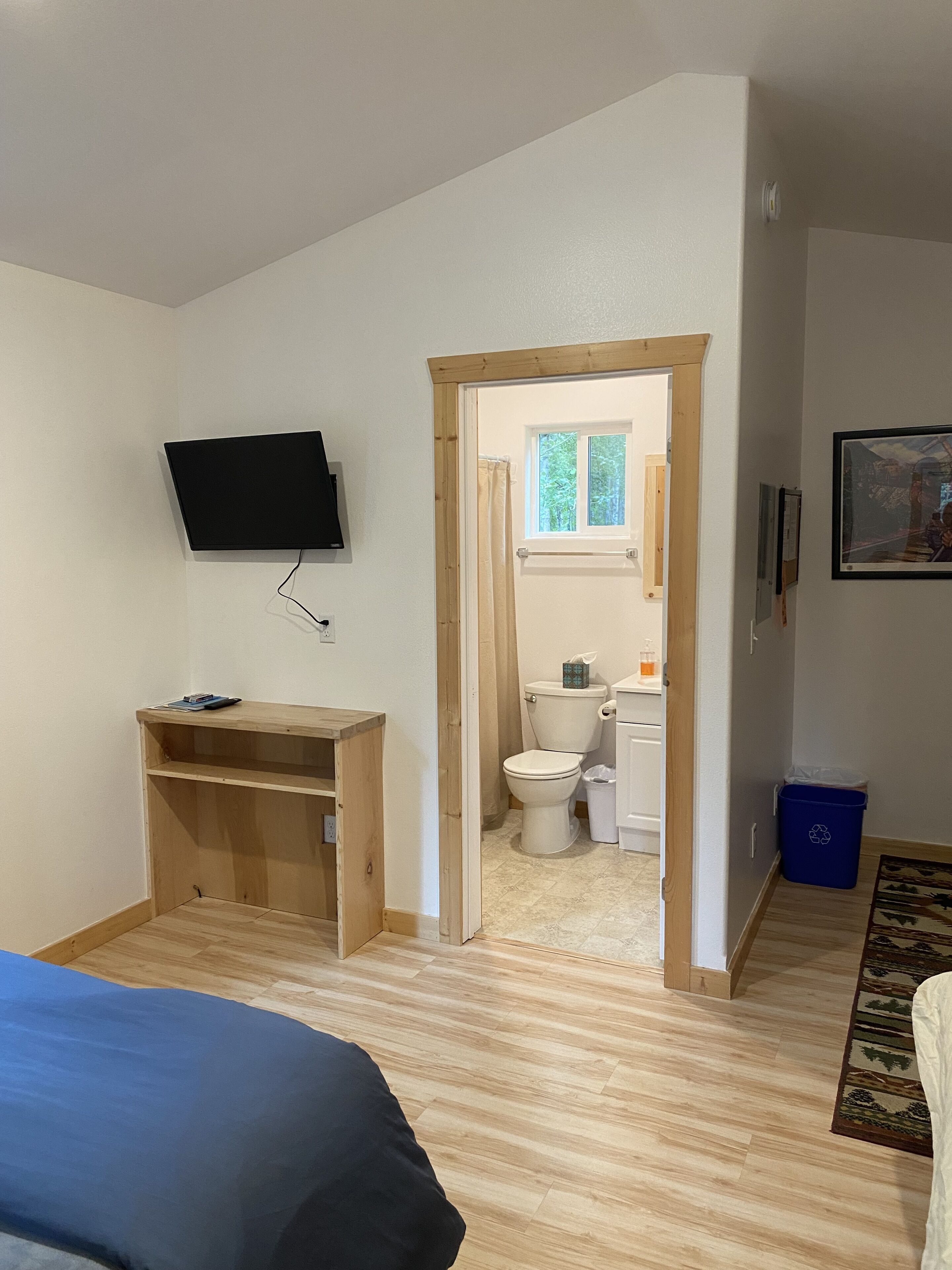 A bedroom with a bed, a TV mounted on the wall, a wooden desk, and a bathroom visible through an open door.