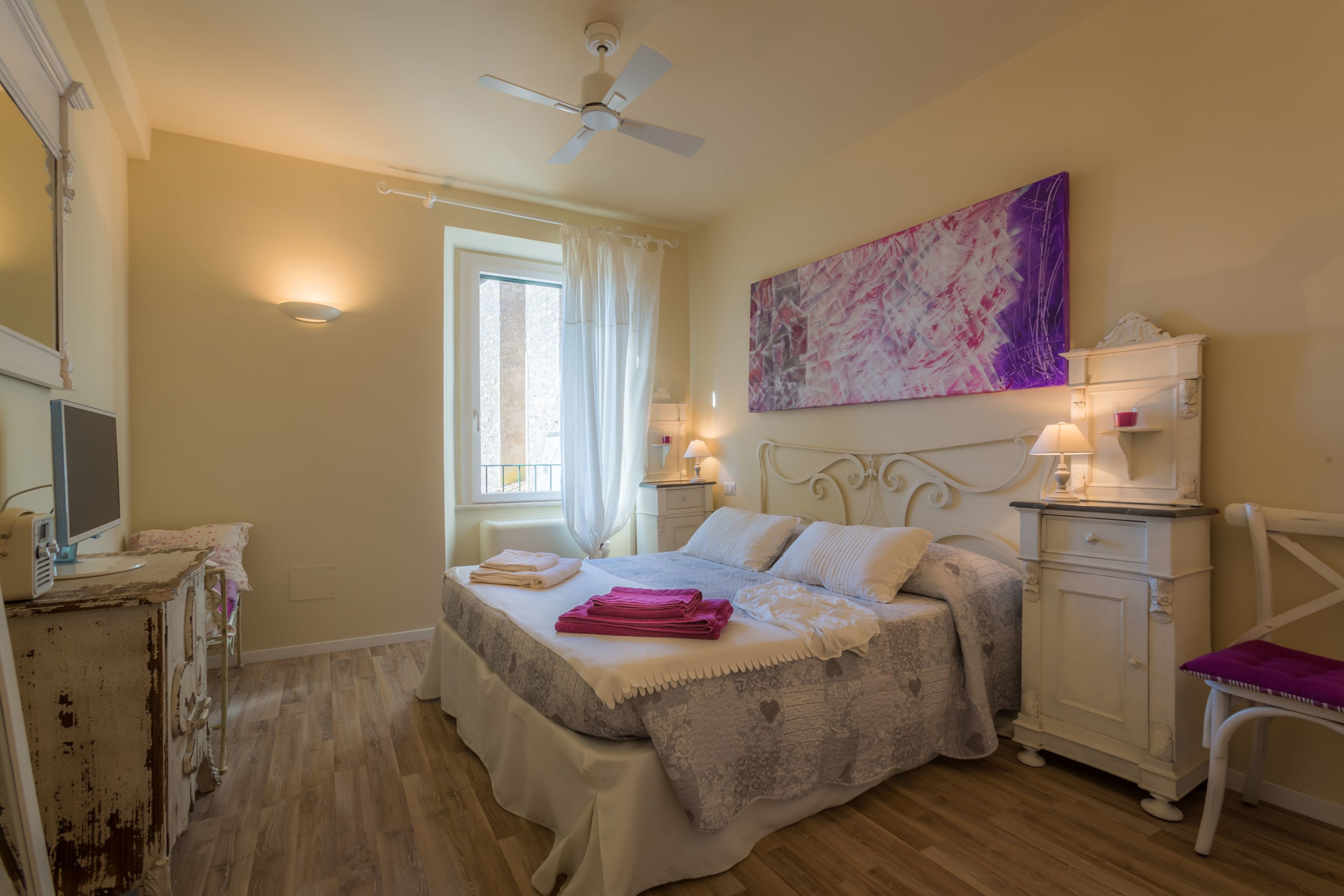 A bedroom with a bed, bedside tables, a chair, a ceiling fan, a window with curtains, and a wall-mounted artwork.