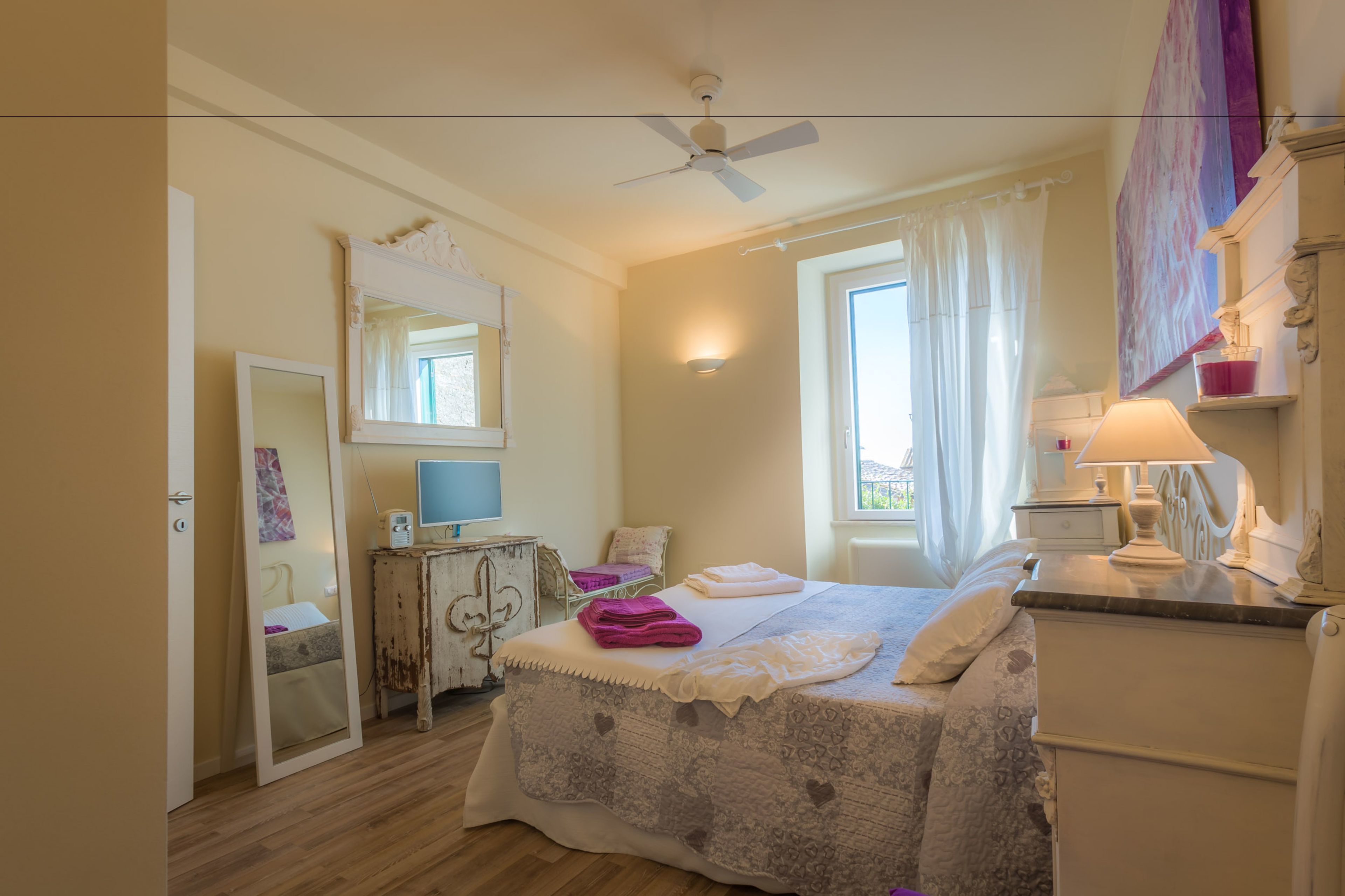 Una camera da letto con un letto, un comodino, uno specchio, una finestra con tende e una ventola a soffitto.