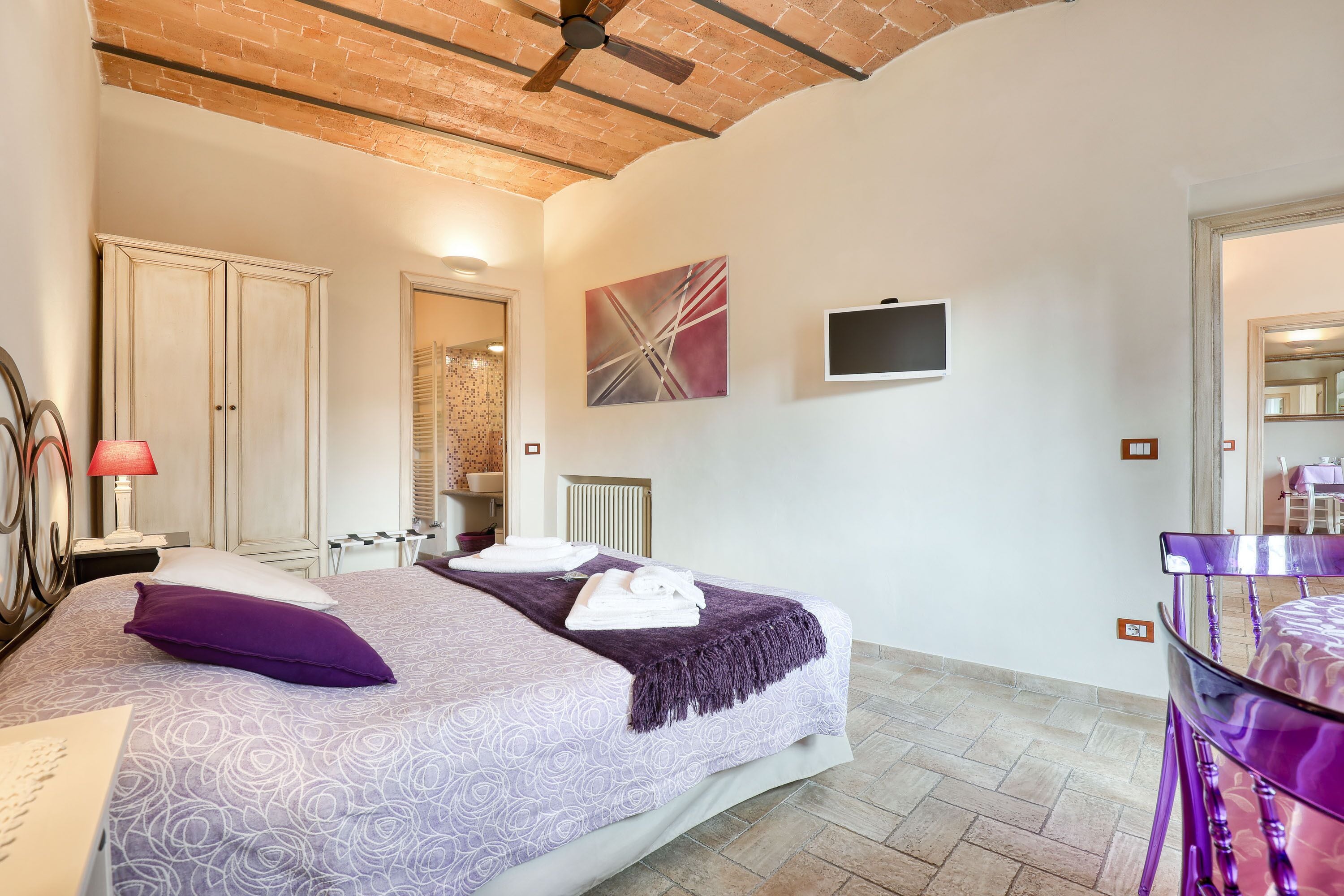 Un dormitorio con cama, televisión, sillón y un baño visible a través de una puerta abierta.