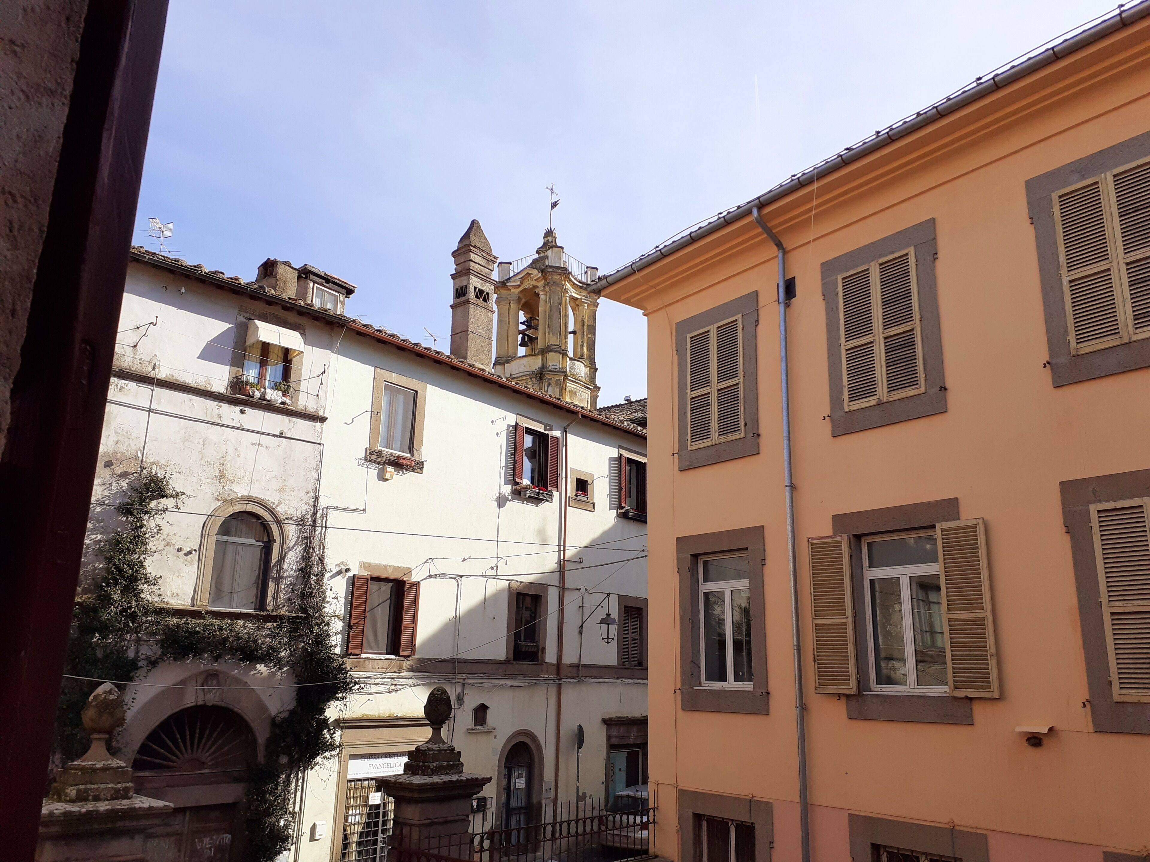 A narrow street with historic buildings, including a church with a bell tower and a building with shutters.