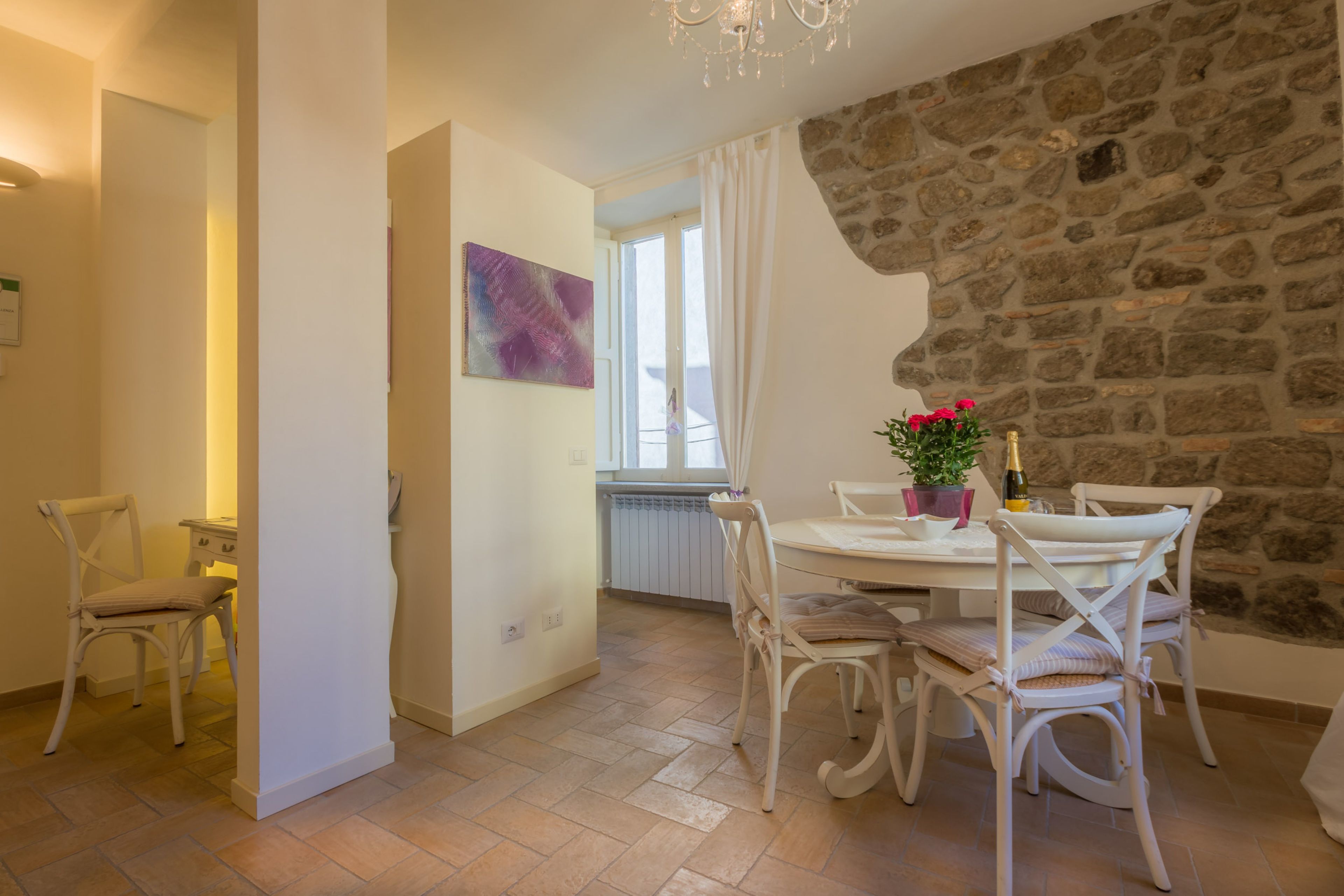 Un comedor con pared de piedra, muebles blancos y una mesa con una botella y flores.