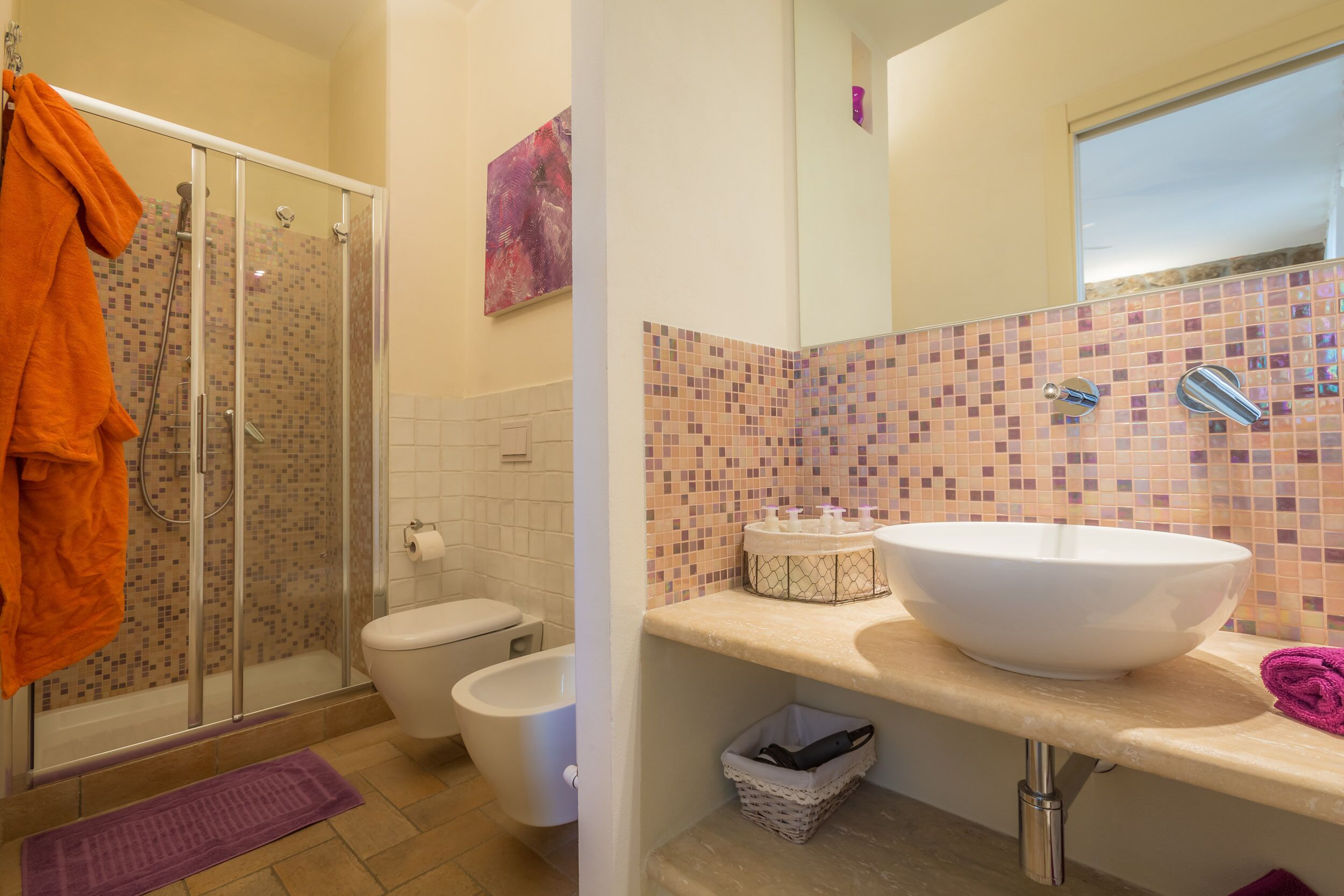 A bathroom with a glass shower enclosure, a toilet, a sink with a mosaic backsplash, and a towel rack with an orange towel.