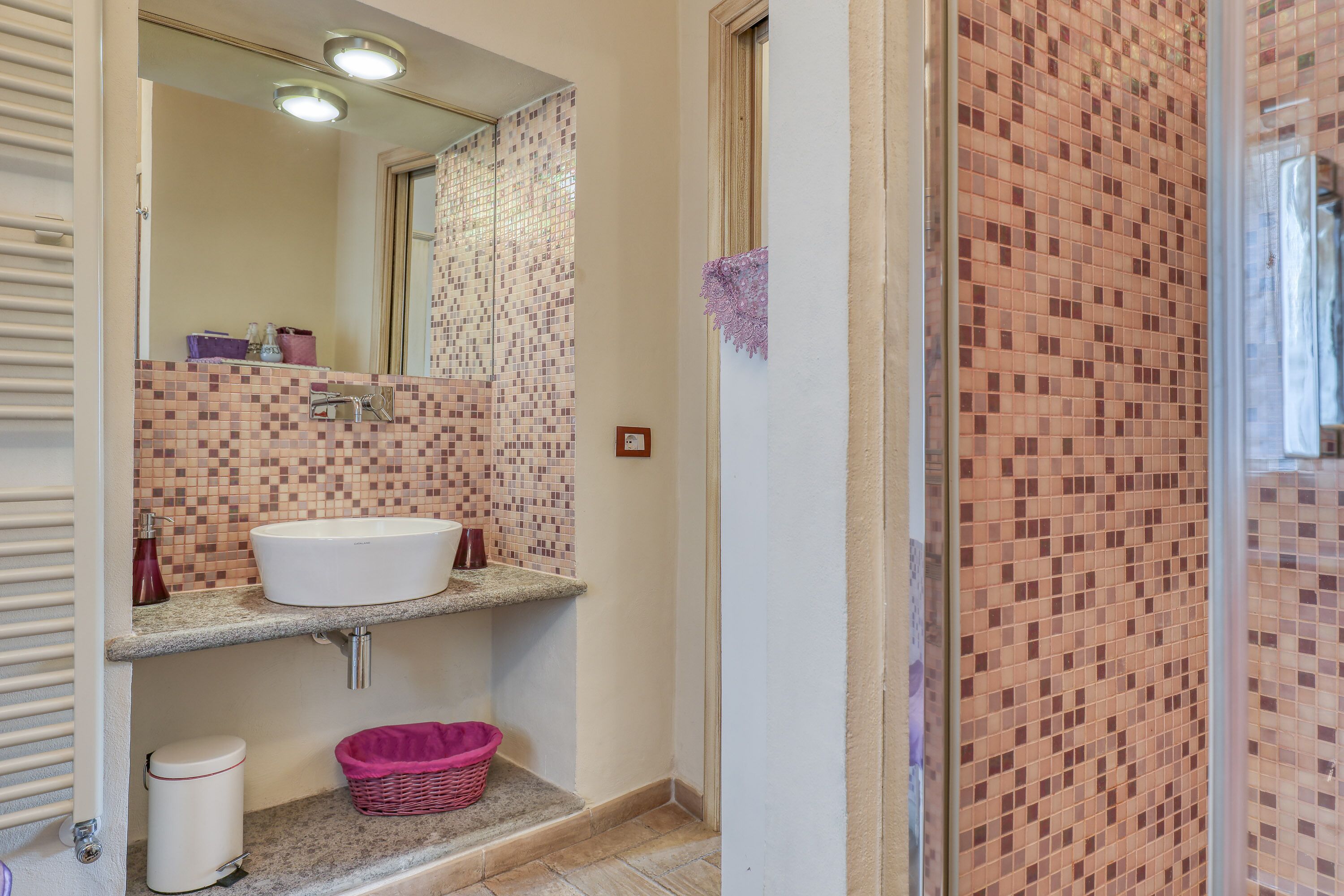 A bathroom with a white sink, a mirror, a tiled shower area, and a pink basket on the counter.