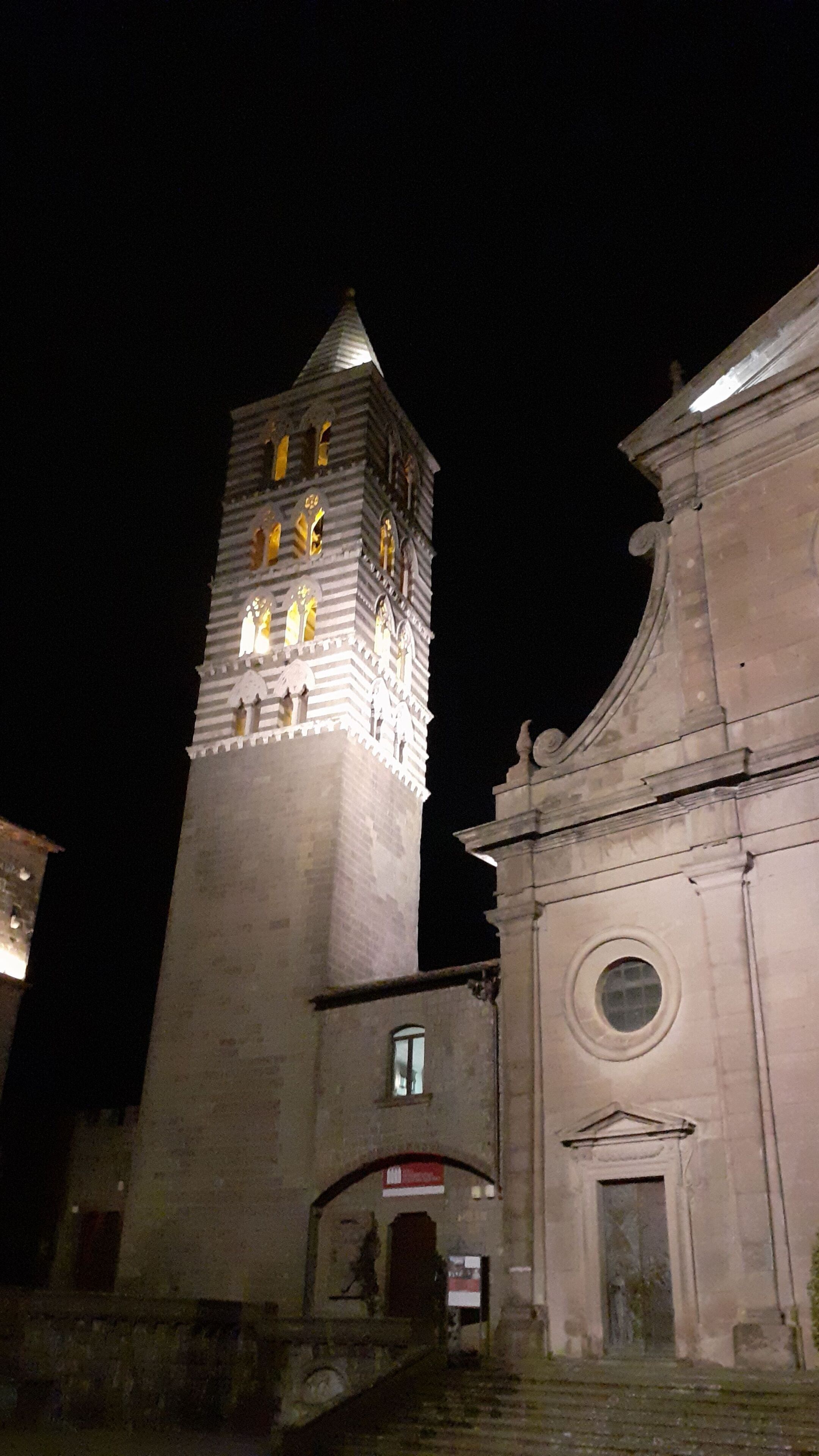 Une tour historique, dotée de fenêtres illuminées et d’une horloge, jouxtant un bâtiment avec une fenêtre circulaire et une porte.