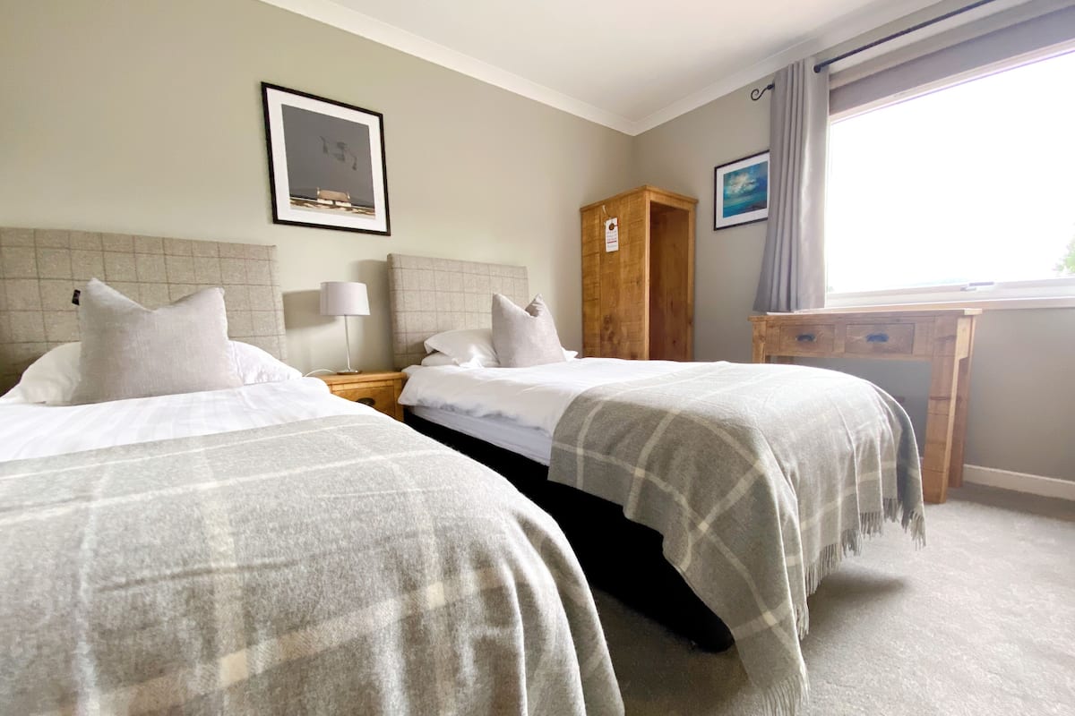 A hotel room with two beds, a wooden wardrobe, a desk with a chair, and a window with curtains.