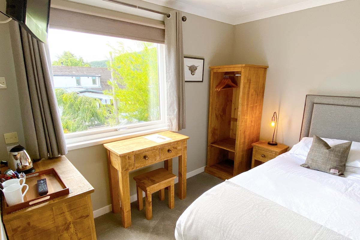 A hotel room with a bed, a wooden desk, a wardrobe, and a window with a view of greenery and buildings.