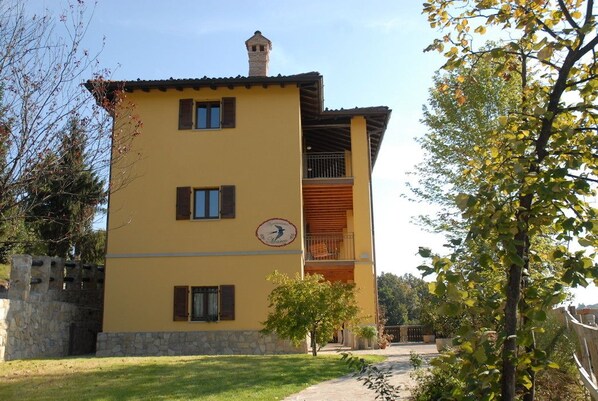 Un bâtiment à deux étages avec une façade jaune, un balcon doté d’une balustrade en bois et un mur de pierre sur le côté gauche.