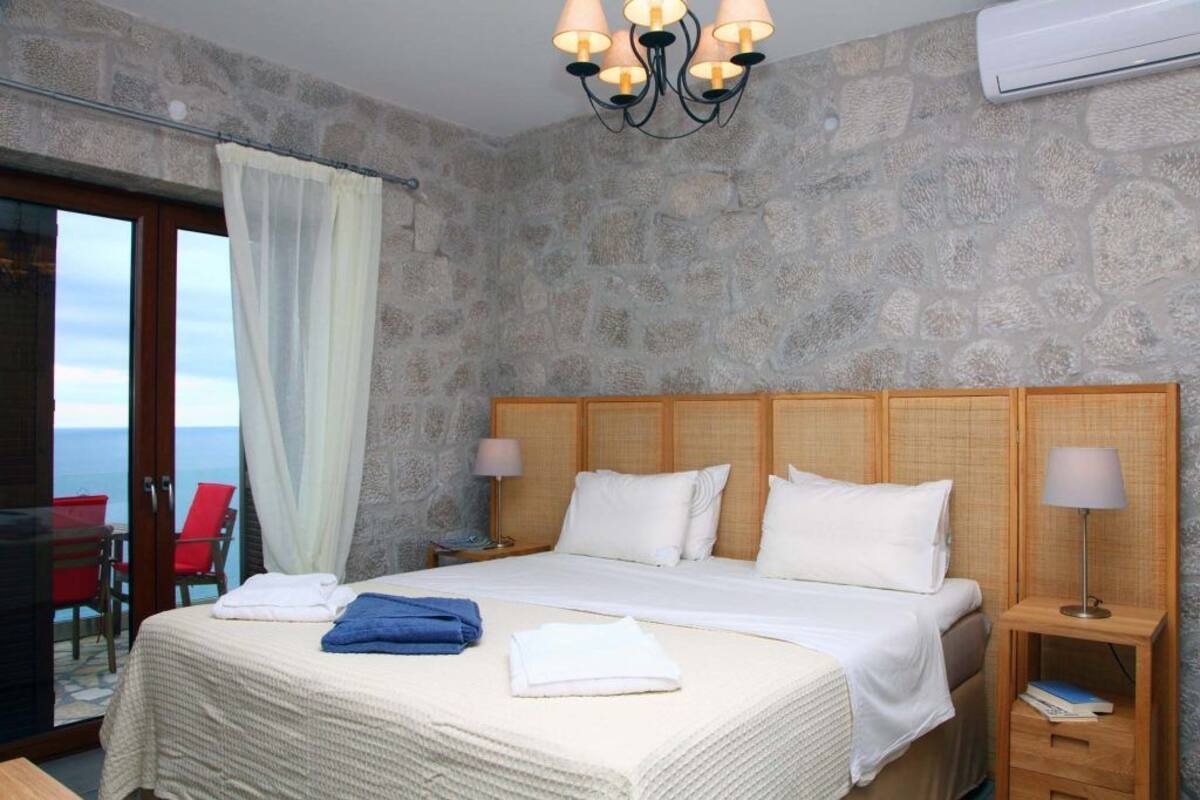 A hotel room with a large bed, a wooden headboard, a nightstand with a lamp, and a view of the sea through a glass door.