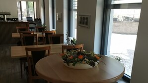 Un comedor con una mesa redonda, sillas y un centro de mesa floral. Hay ventanas grandes con persianas, una zona de cocina al fondo y suelo de madera.