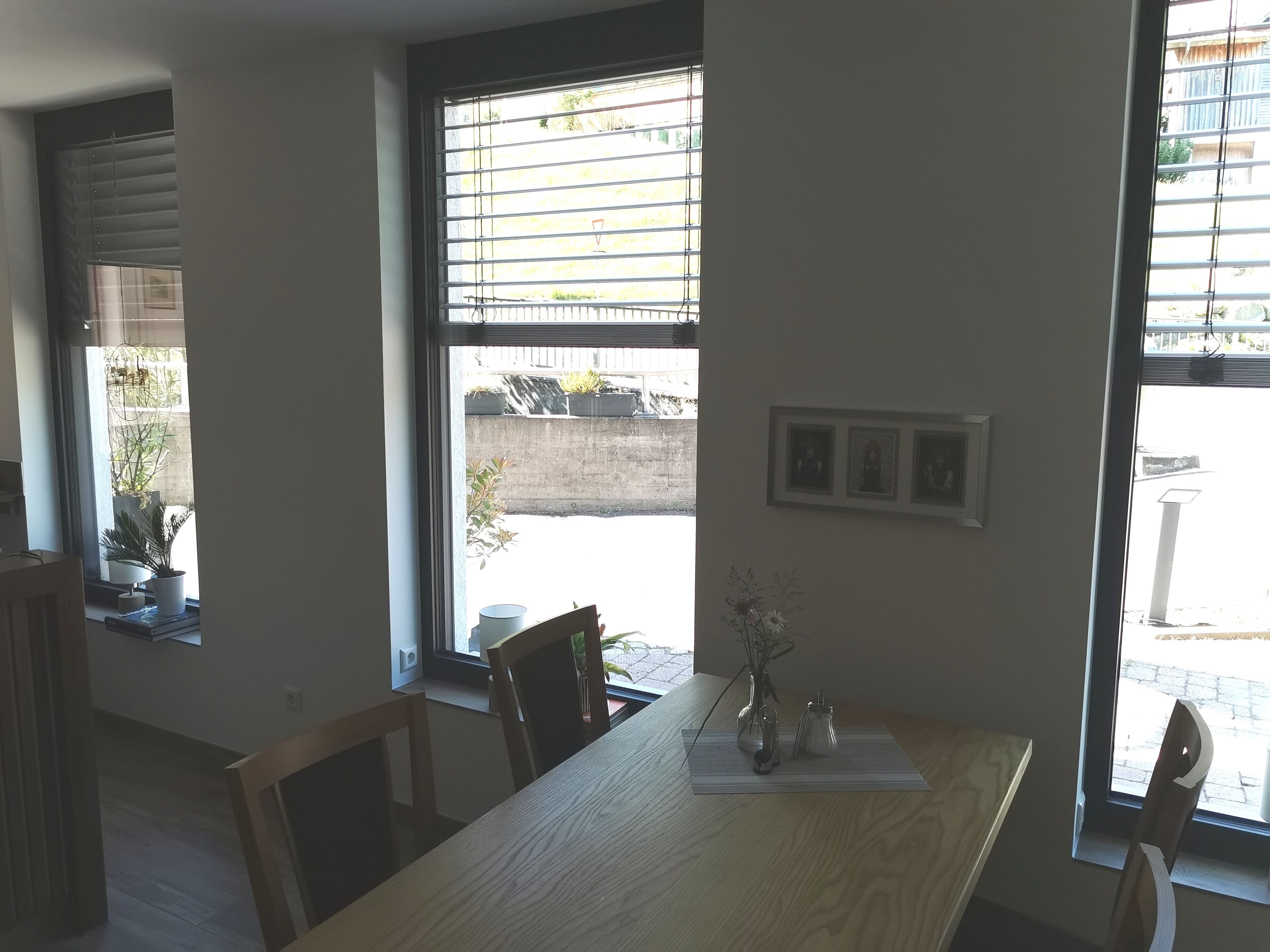 A room with a wooden table, chairs, a window with blinds, and a wall-mounted picture frame.