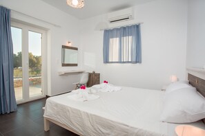 A hotel room with a large bed, a desk, a chair, a mirror, and a window with curtains. - Villa Maria (Paros)