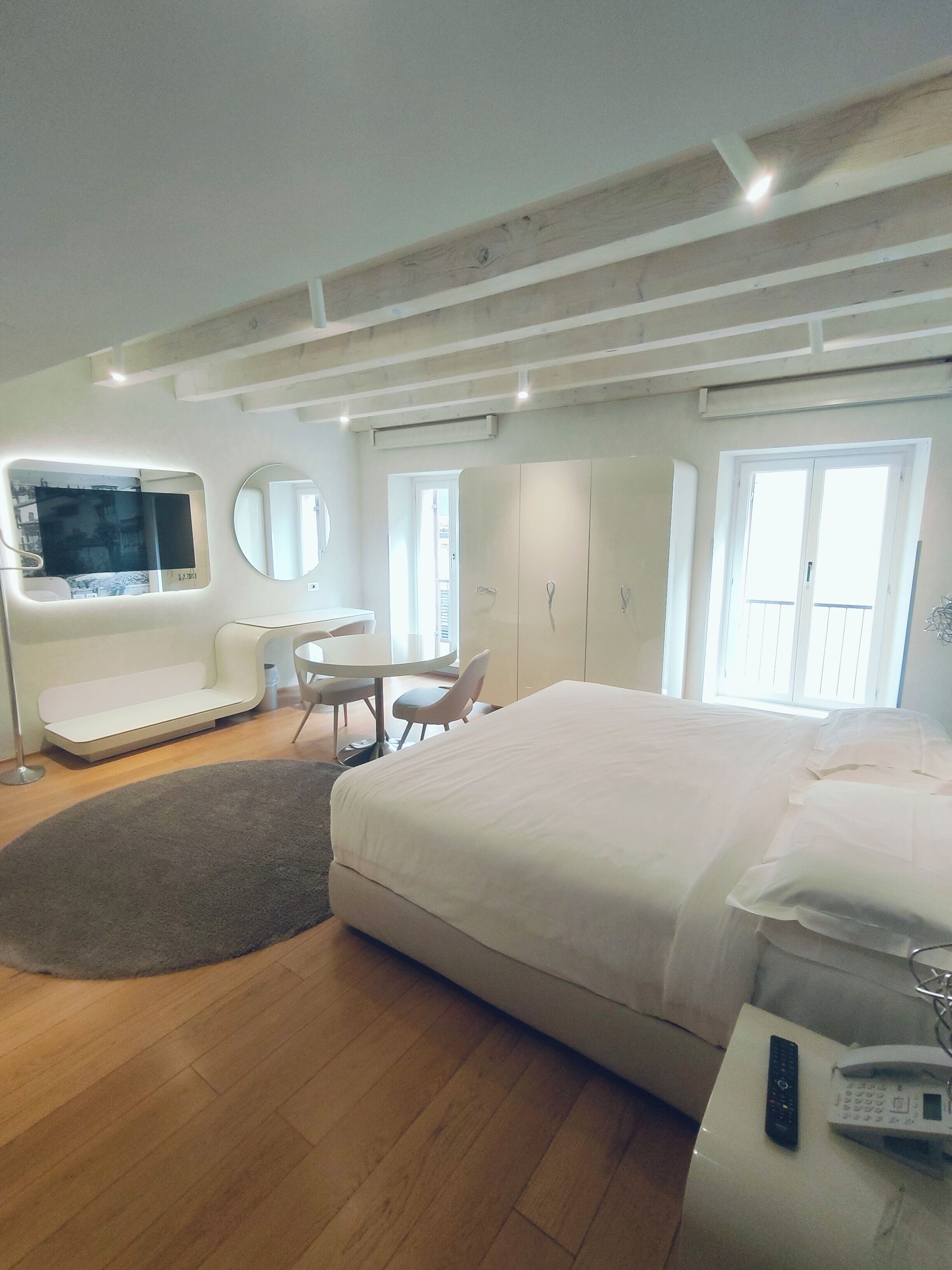 A modern hotel room with a large bed, a desk, a round mirror, and a TV mounted on the wall.