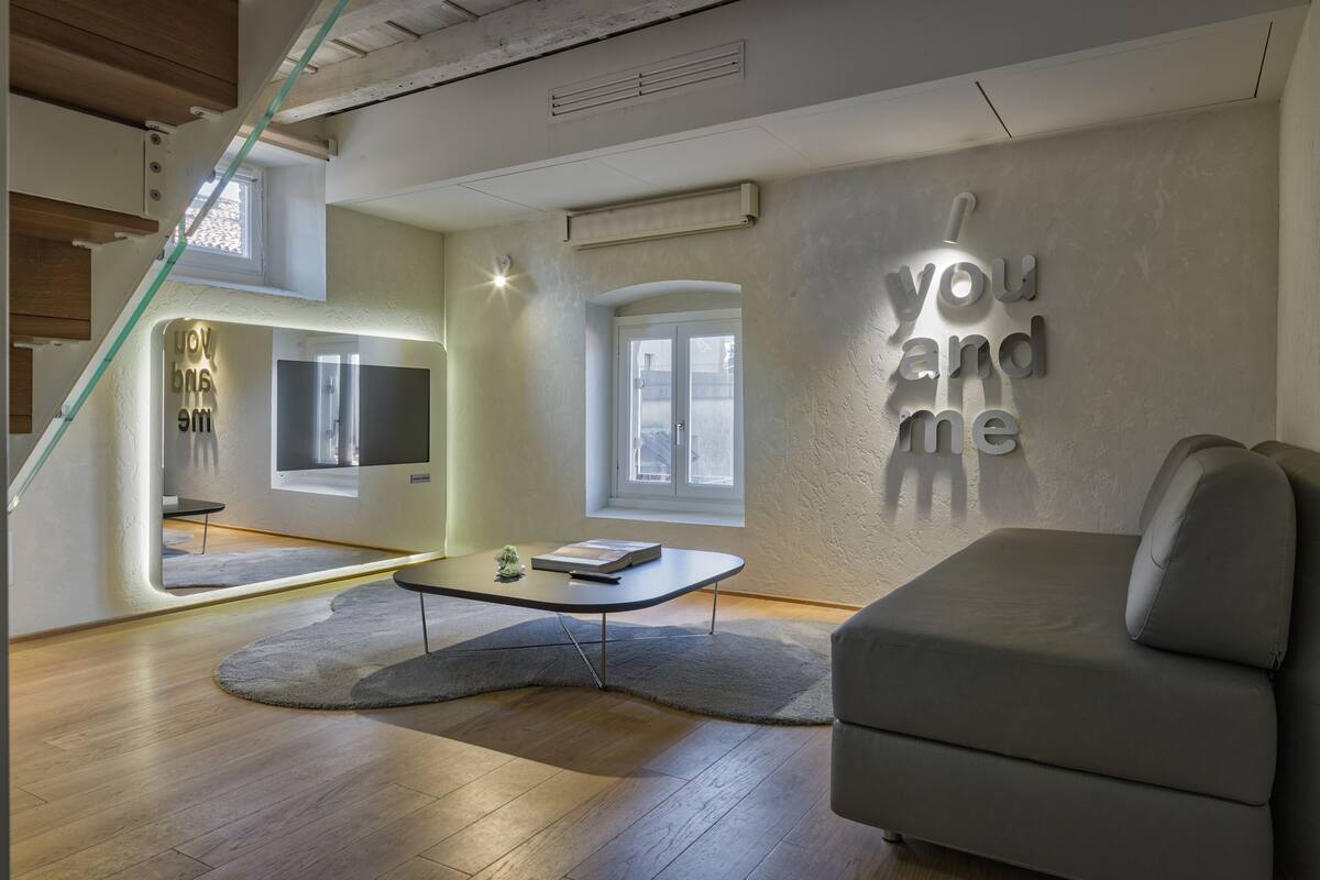 A modern living room with a grey sofa, a small table, and a TV mounted on the wall. There is a window with a view of the outside, and a sign on the wall that reads "you and me".