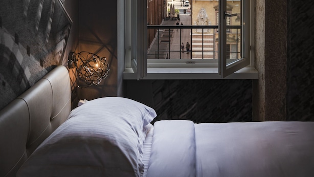 Une chambre à coucher avec un lit, une table de chevet, une lampe et une fenêtre donnant sur des immeubles.