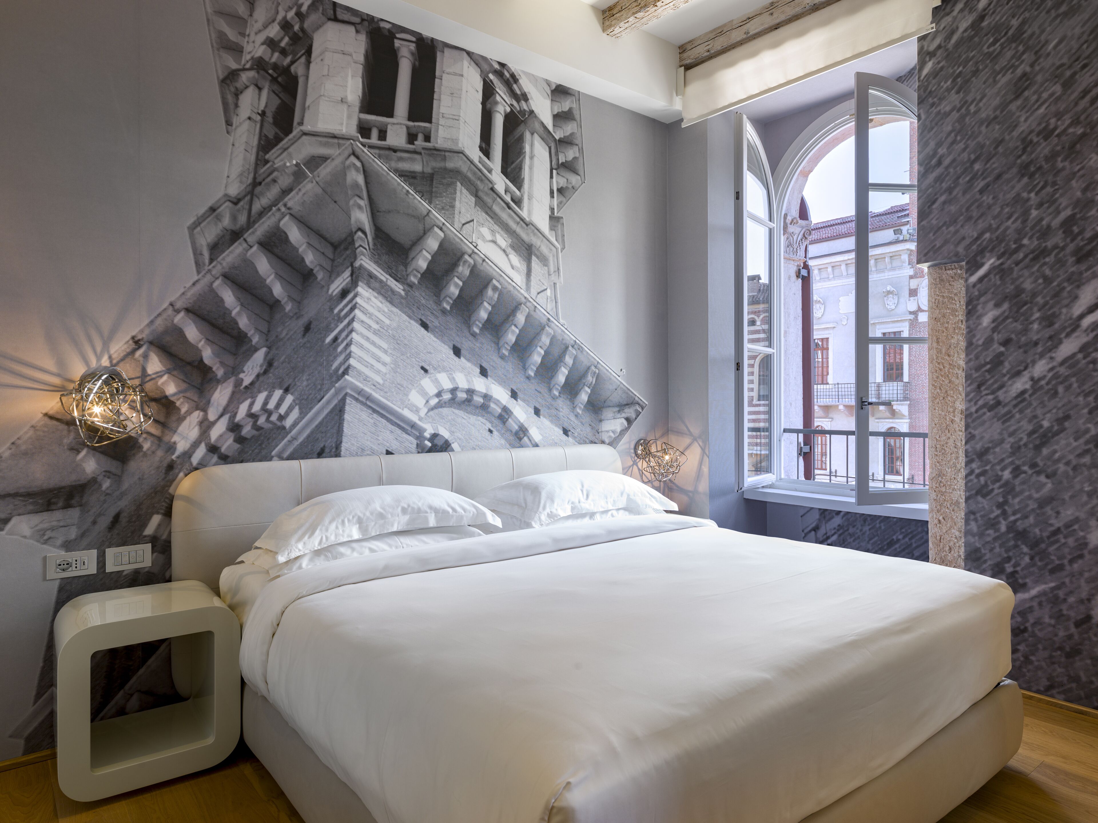 Un dormitorio con una cama grande, una lámpara moderna y vistas a edificios a través de una ventana.