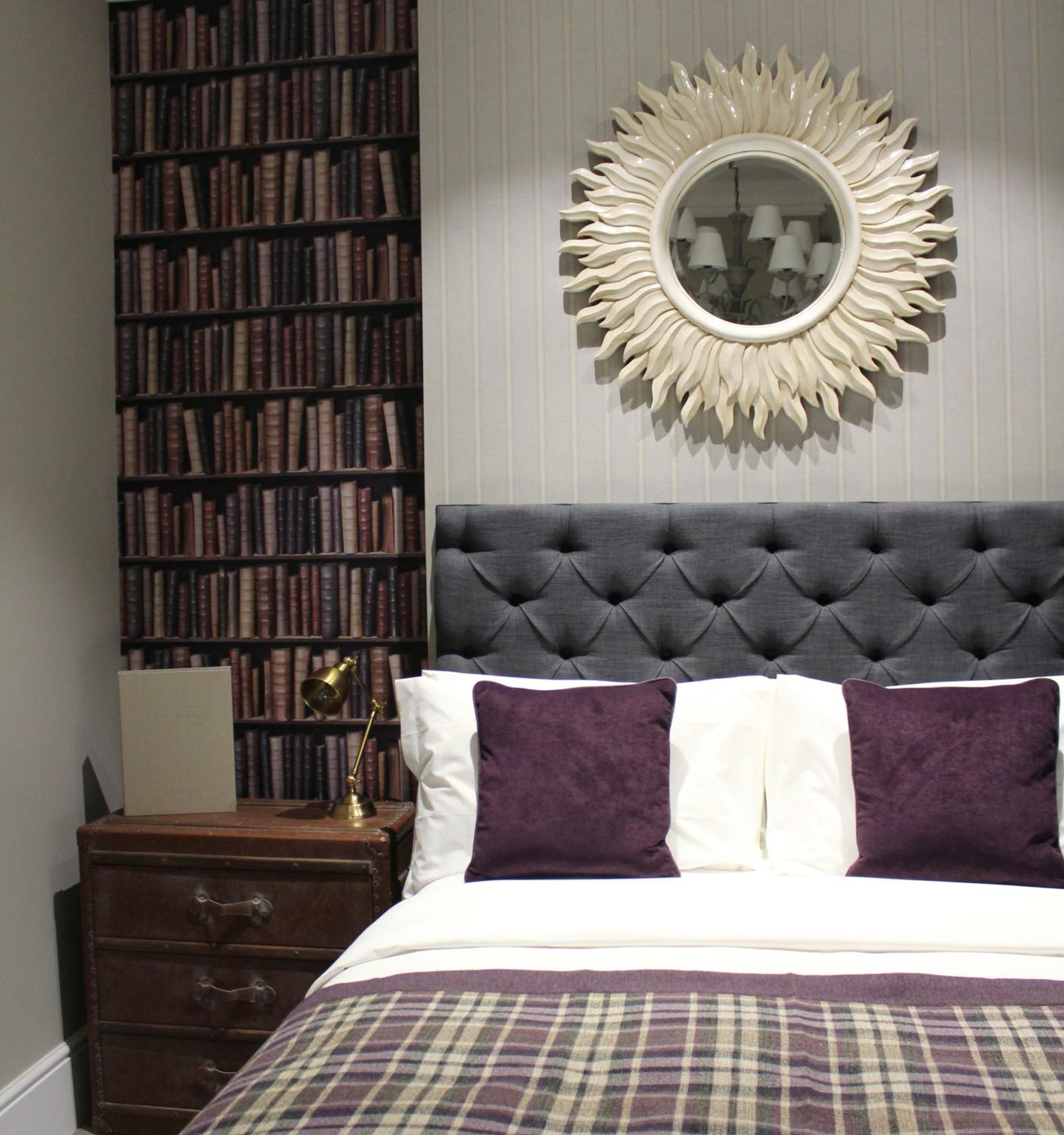 A bedroom with a tufted headboard, a large mirror with a sunburst frame, a bookshelf filled with books, and a plaid bedspread.