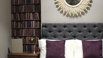 A bedroom with a tufted headboard, a large mirror with a sunburst frame, a bookshelf filled with books, and a plaid bedspread.