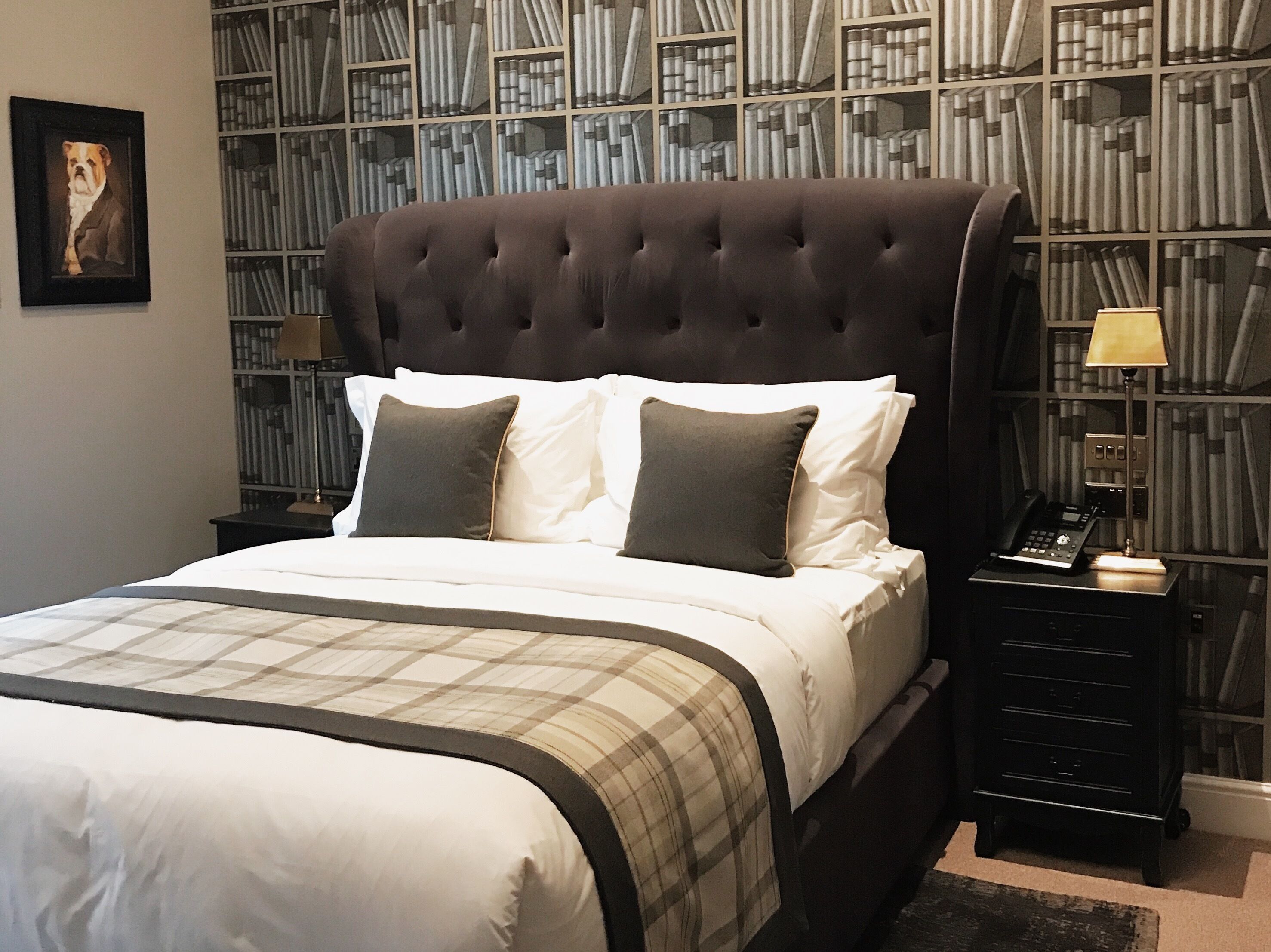 Ein Bett mit dunklem Kopfteil, weißer Bettwäsche und einer karierten Decke. Ein Nachttisch mit Lampe und Telefon steht daneben. Im Hintergrund ist ein Bücherregal mit Büchern zu sehen.