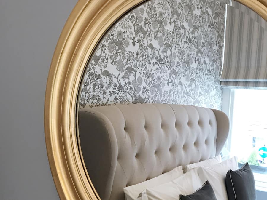 A round mirror with a gold frame reflecting a room with a tufted headboard sofa, white and dark gray pillows, and a floral wallpaper background.