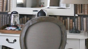Camera d'albergo con un ampio specchio, una scrivania con bollitore, una lampada e una sedia. Si nota una libreria con libri e un quadro incorniciato appeso al muro.