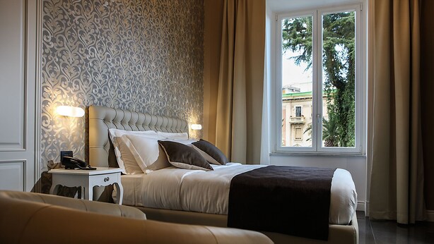 A hotel bedroom with a bed, bedside table, lamp, and a window with curtains. - Villa San Martino (Naples)