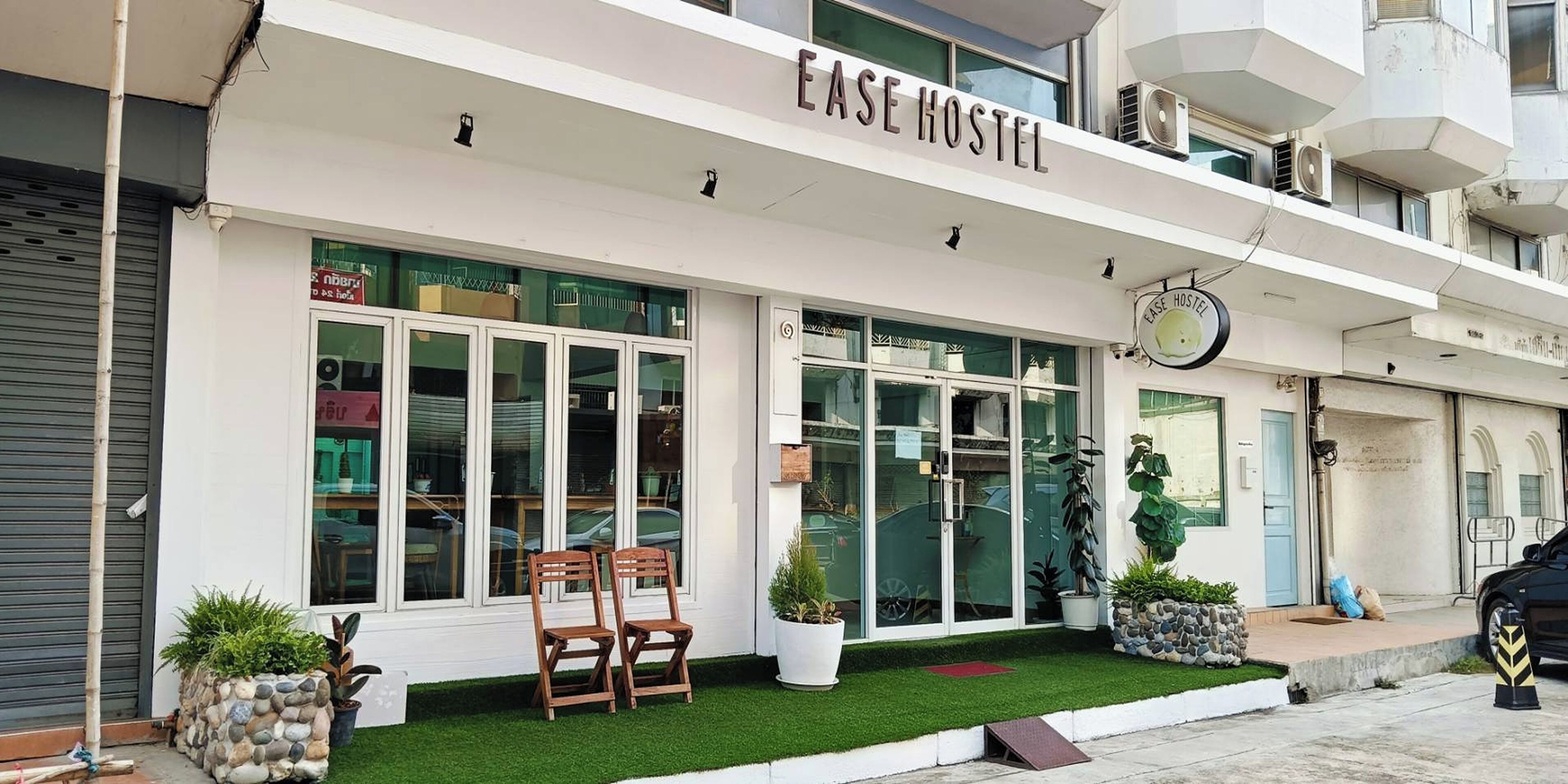 A hostel named "EASE HOSTEL" with a modern facade, large windows, and a small outdoor seating area with chairs and potted plants.