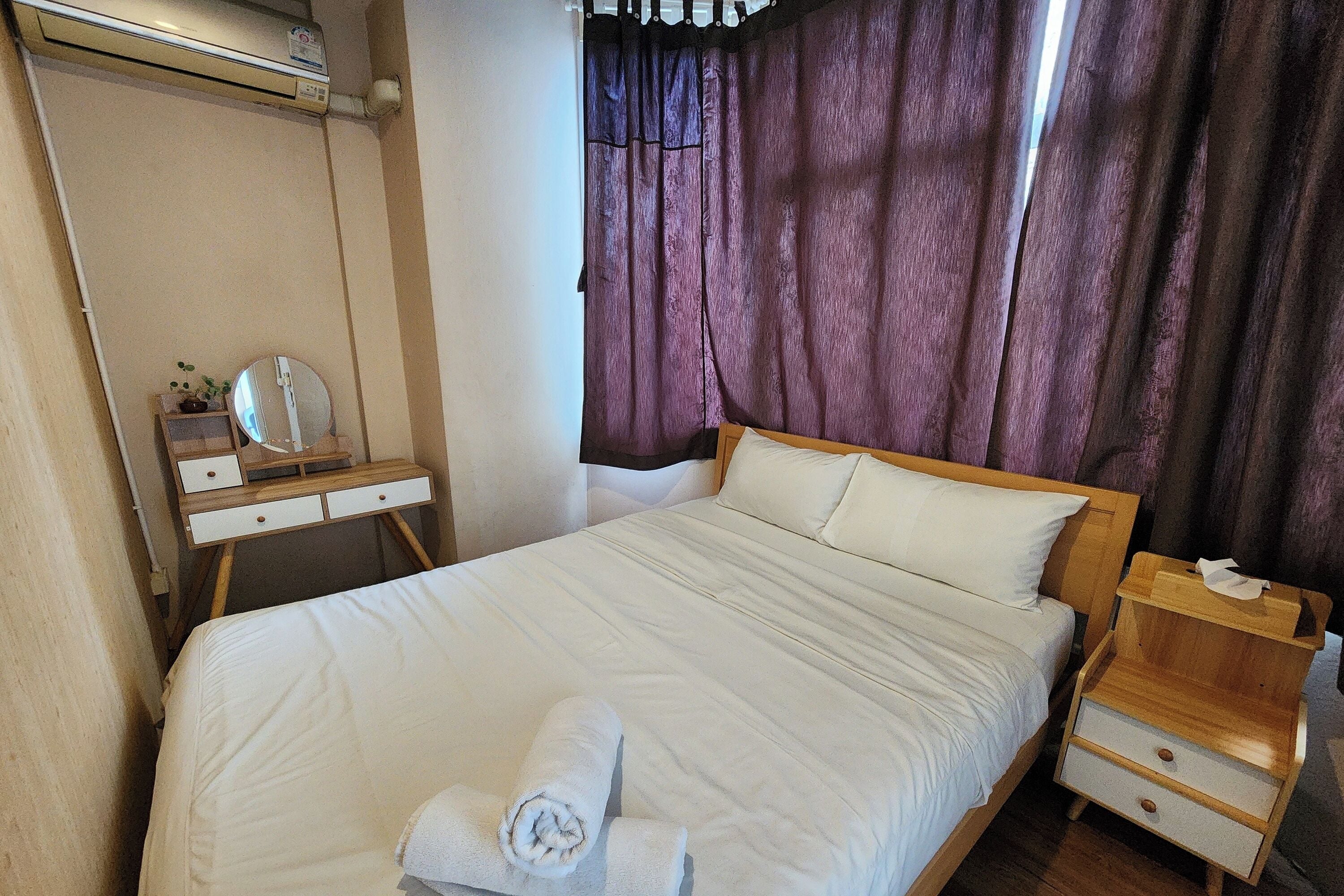 A neatly made bed with a white comforter and two pillows, a wooden nightstand with a tissue box, a small wooden dresser with a mirror, and a window with purple curtains.