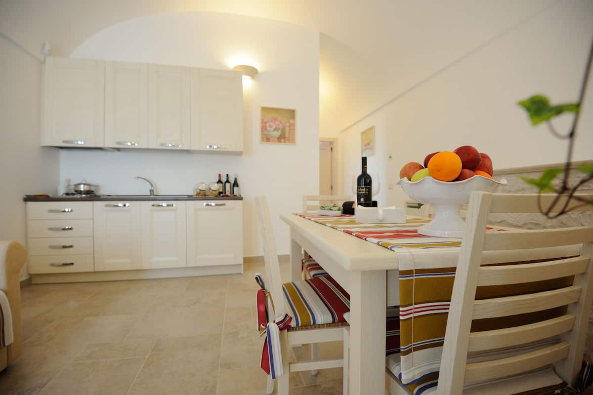 Une cuisine avec des meubles blancs, une table à manger recouverte d’une nappe à rayures et un saladier rempli de fruits.
