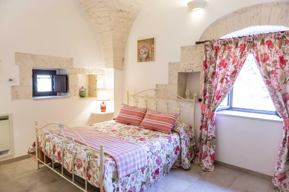 Une chambre à coucher avec un lit, une fenêtre ornée de rideaux à motifs floraux, un téléviseur fixé au mur et une petite étagère avec une lampe.