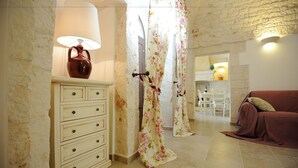 A room with a wooden cabinet, a lamp, floral curtains, a sofa, and a dining area in the background.