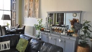 A living room with a leather couch, a dining table with chairs, a cabinet with jars and a sign, and a large window. - The Bank Guest House (Hawick)