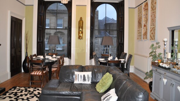 A living room with a leather sofa, dining table, chairs, and a chandelier. - The Bank Guest House (Hawick)