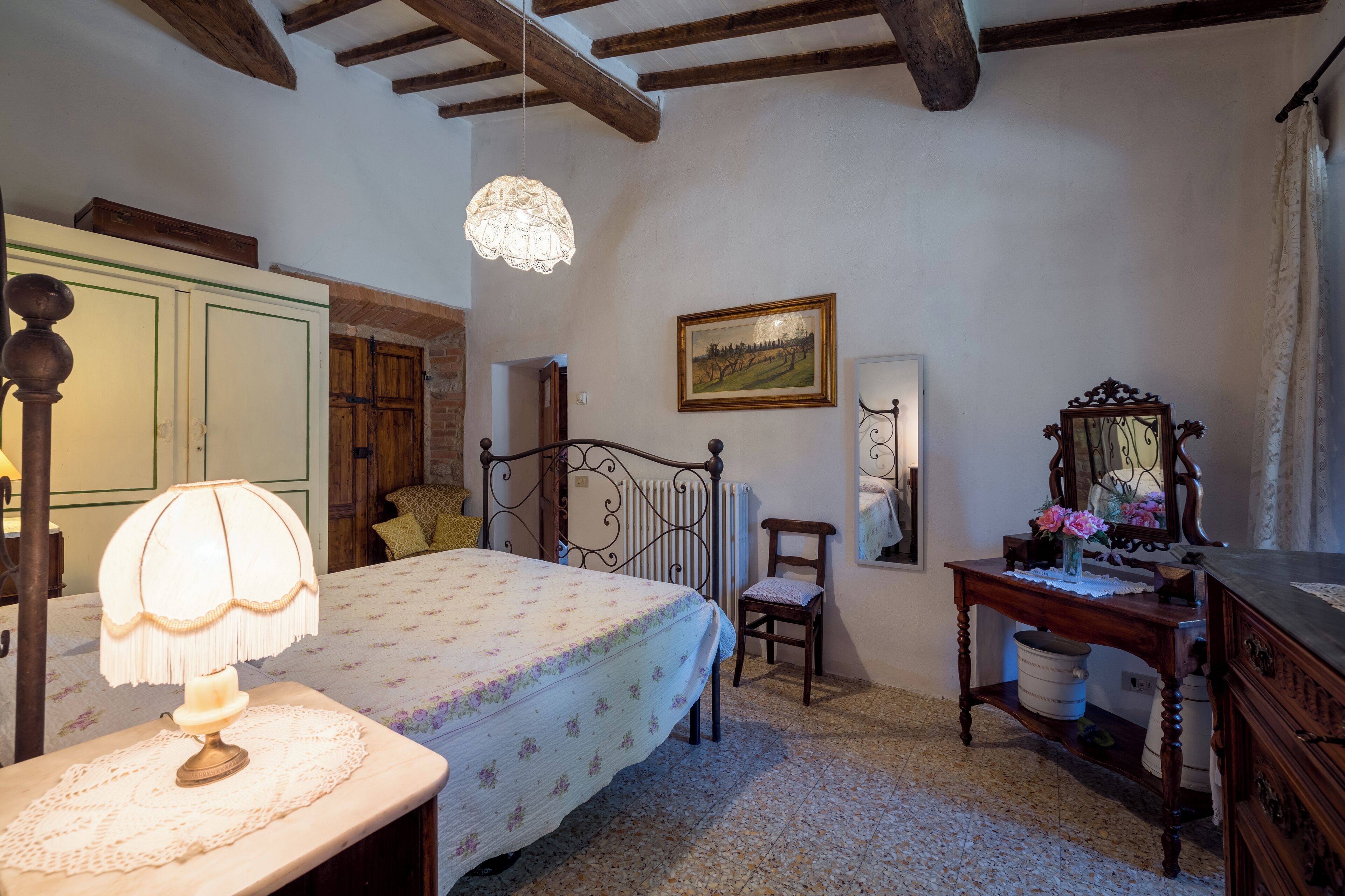 A bedroom with a bed, a bedside table with a lamp, a wooden chair, a dresser, a mirror, and a framed picture on the wall.