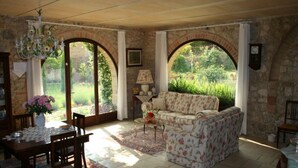 Ein gemütliches Wohnzimmer mit einer blumengemusterten Couch, einem Esstisch mit Stühlen und einer Deckenleuchte. Zwei große Bogenfenster bieten Blick auf einen Garten.