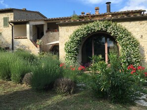 Un bâtiment rustique en pierre, doté d’un patio couvert, d’une grande fenêtre en arc de cercle et d’un jardin où poussent diverses plantes et fleurs.